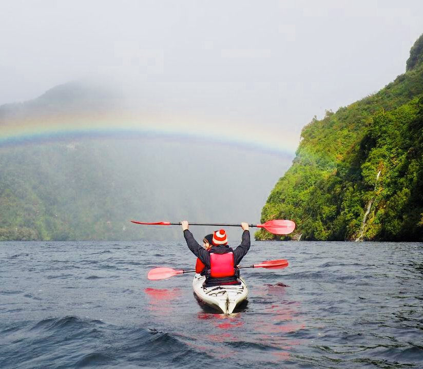 Two kayakers in double kayak with a rainbow above them. The person in the rear seat is holding their paddle in the air. 